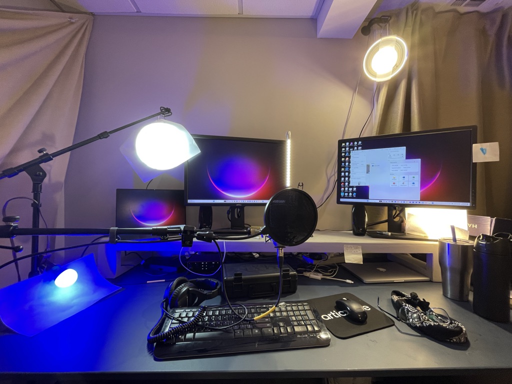 Image of a laptop with two external monitors, a wireless keyboard a wireless mouse, and 4 lightbulbs of varying color placed around the setup facing the camera.